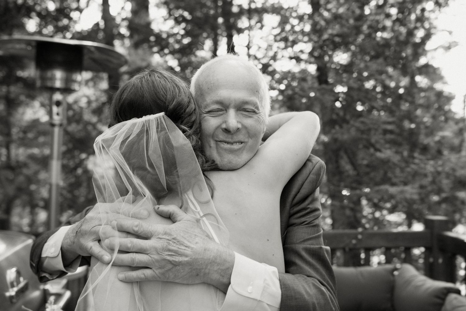 Emotional black and white photo of bride hugging smiling bald man at outdoor wedding.