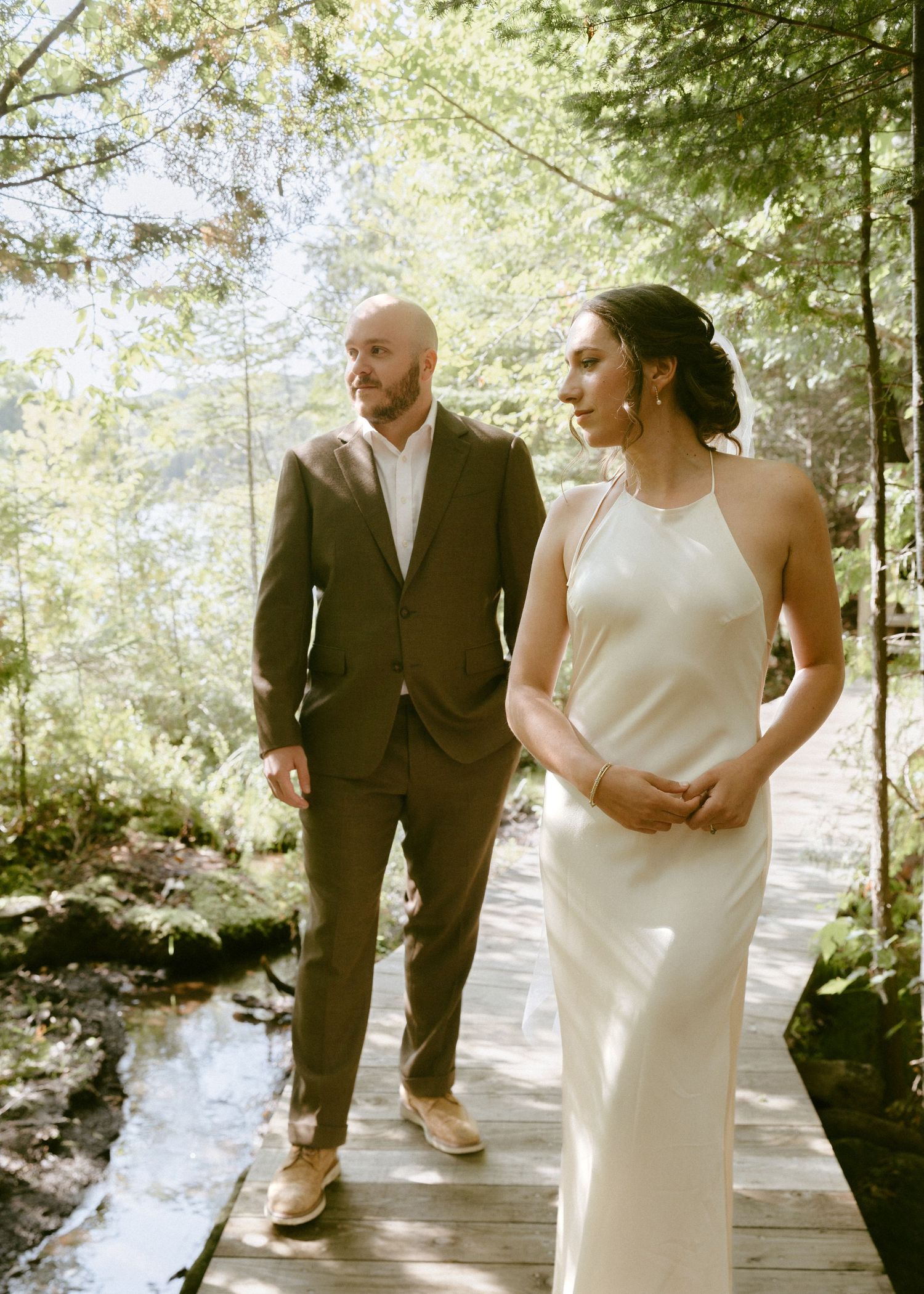 Bride in sleek satin halter gown and groom in brown suit walk along a wooden bridge in sunlit woods.
