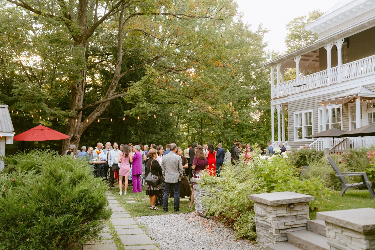Wedding reception guests gather along a garden pathway beside a large historic home surrounded by trees.