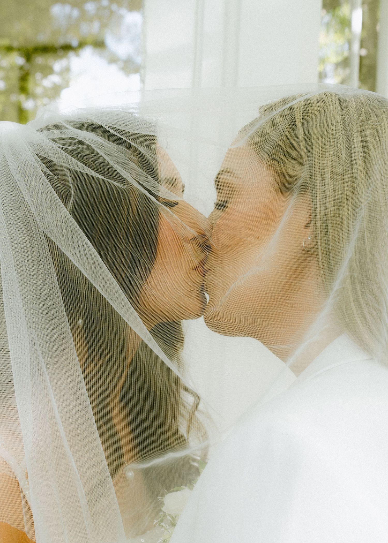 Two brides kissing under a sheer wedding veil in a romantic close-up photo.
