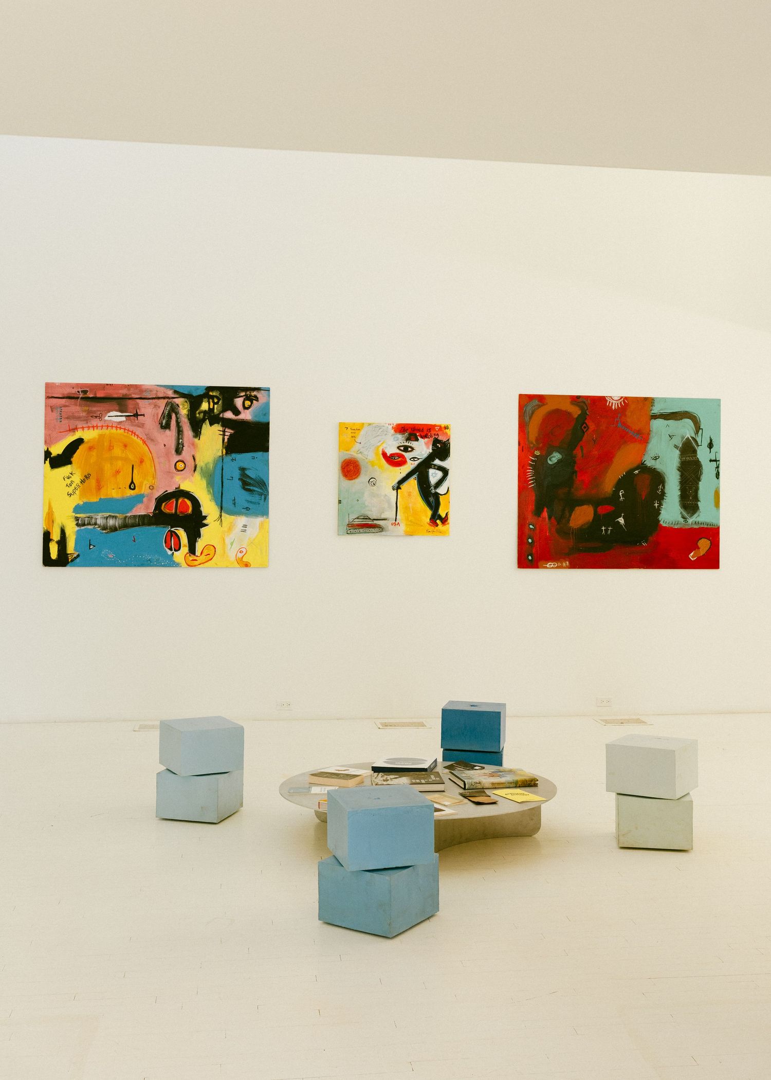 Modern art gallery interior with three colorful abstract paintings and light blue cube seating arrangements on display.