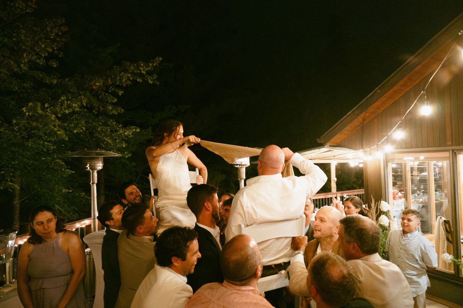 Wedding guests lifting bride and groom on chairs during festive outdoor reception.