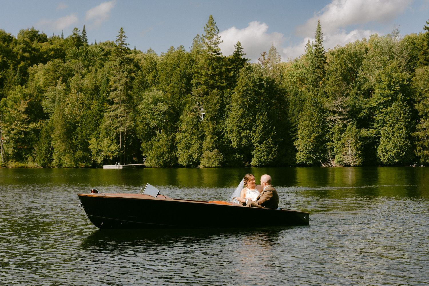 Newlyweds sit close together in a dark wooden motorboat gliding across a peaceful forested lake.