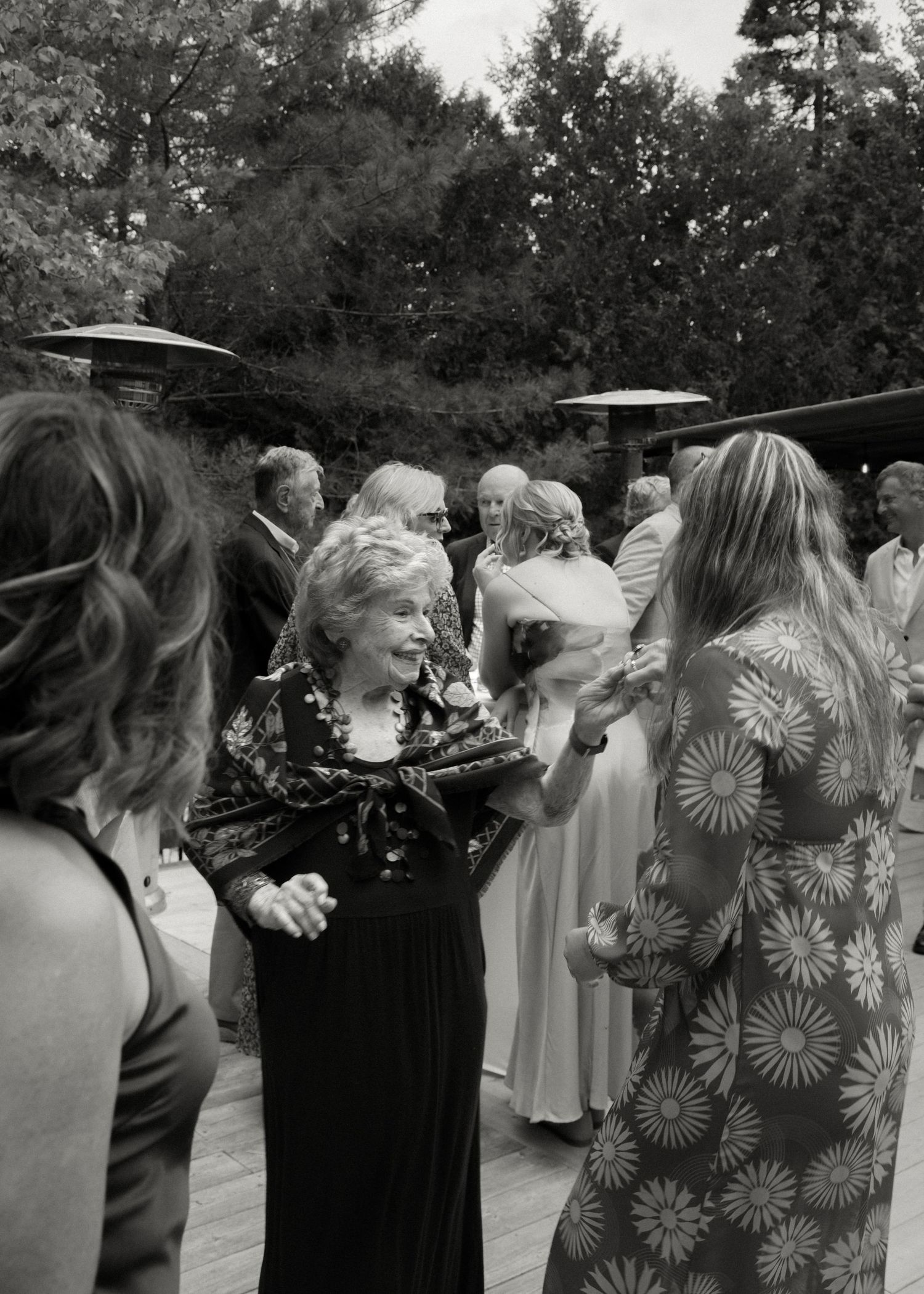 Black and white photo of multigenerational wedding guests mingling outdoors under patio umbrellas.