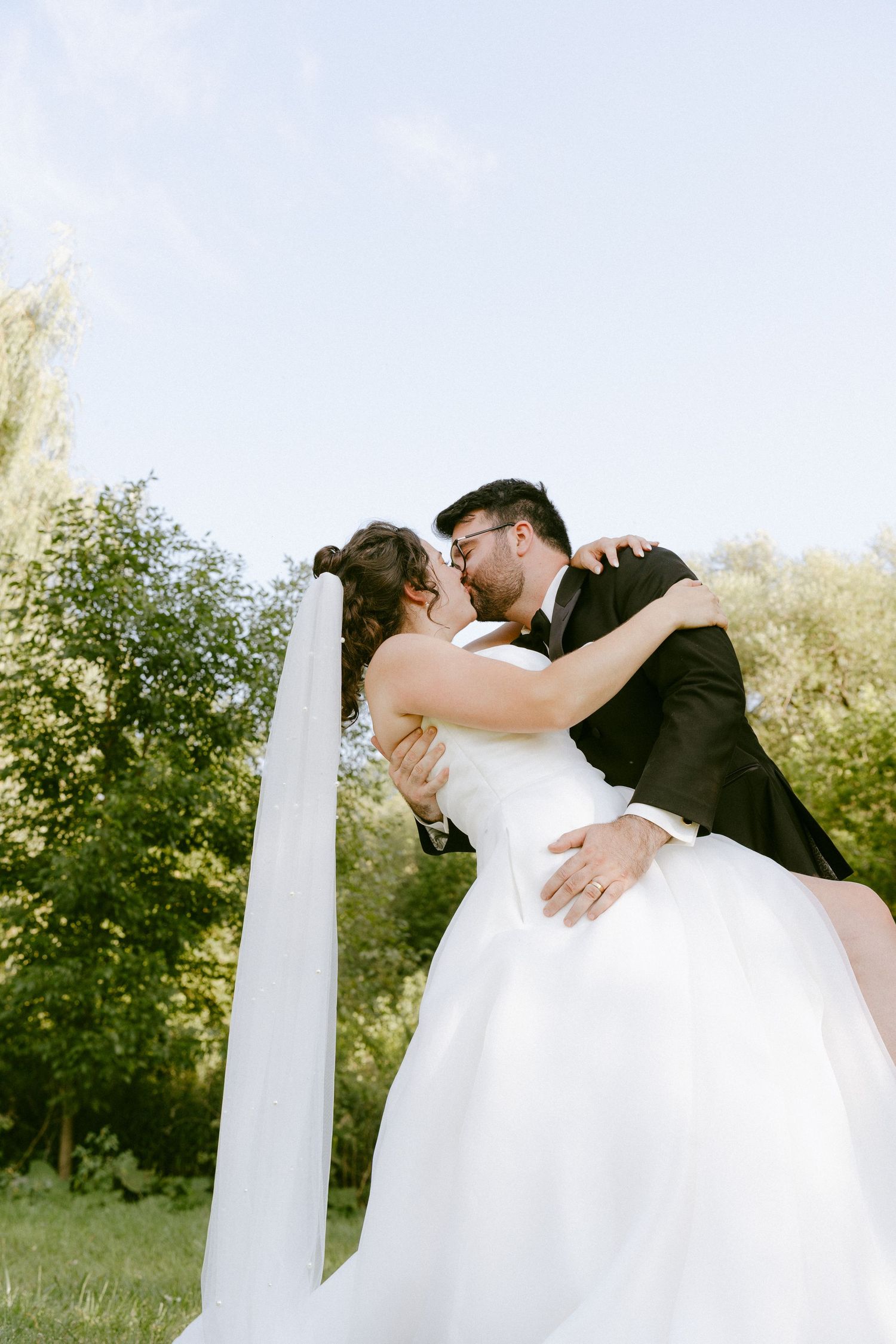 Groom lifts and kisses bride in white gown and veil against a bright blue sky outdoors.