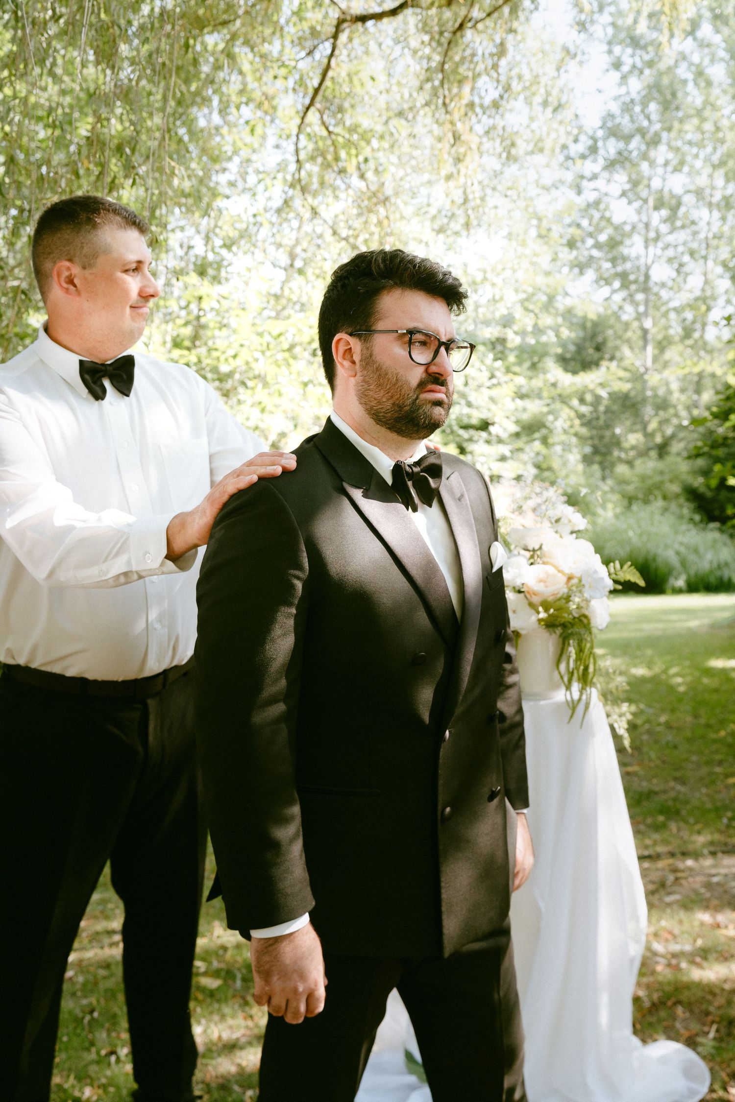 Groomsman adjusts groom's black tuxedo jacket before outdoor wedding ceremony.