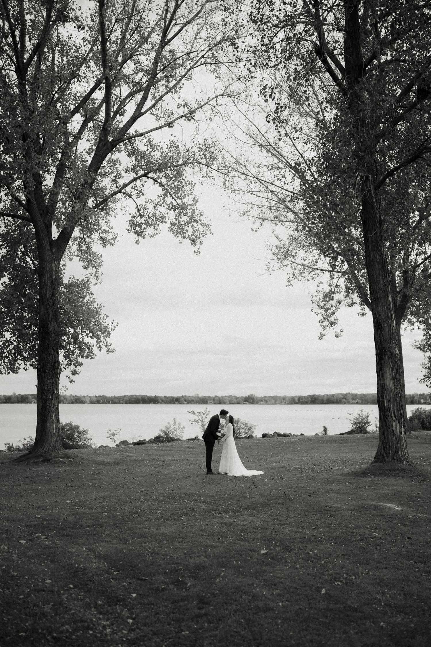 Black and white photo of a bride and groom embracing between tall trees by a lake on their wedding day.