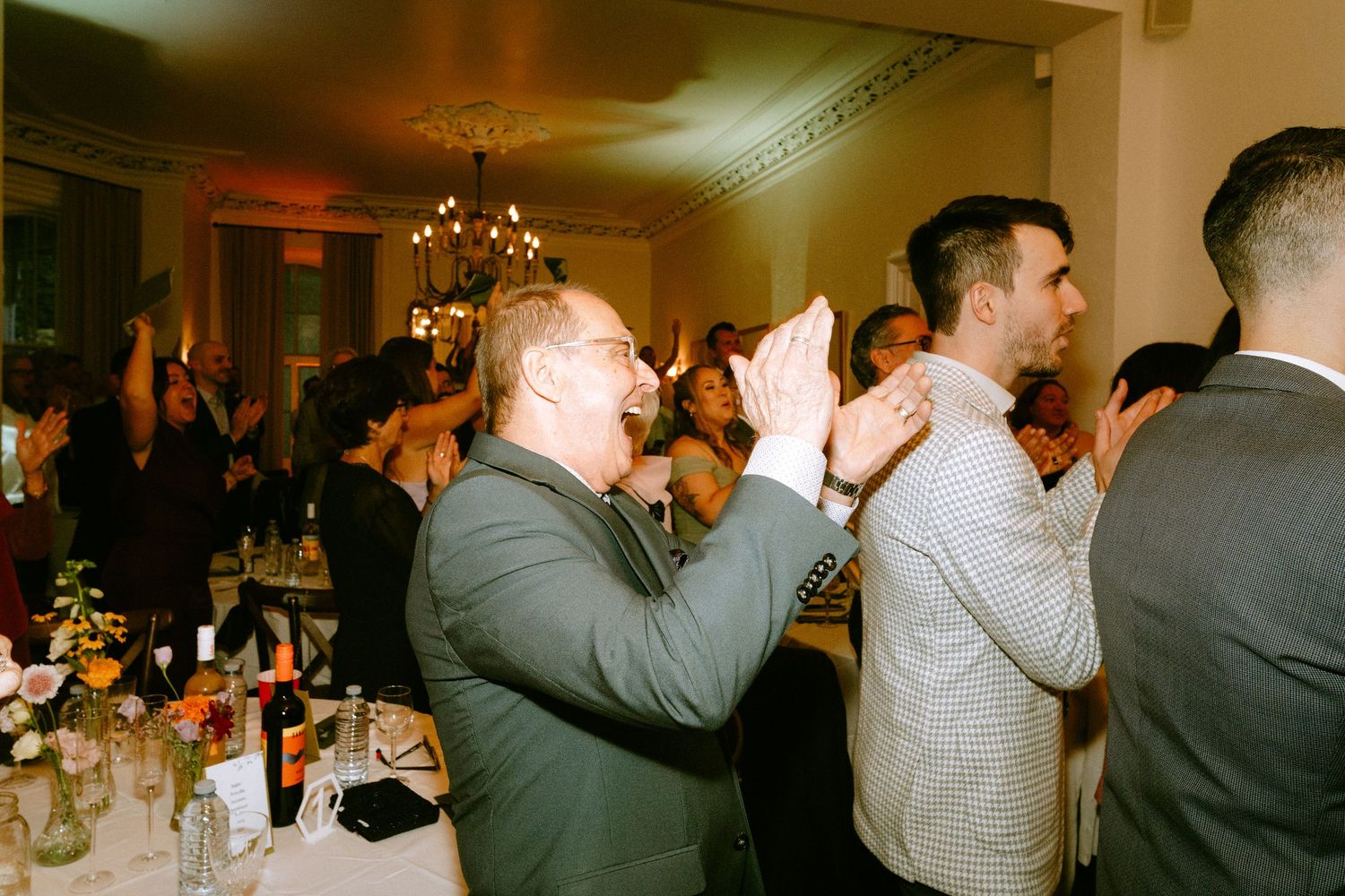 Wedding guests clapping and celebrating at indoor reception dinner with chandelier lighting.
