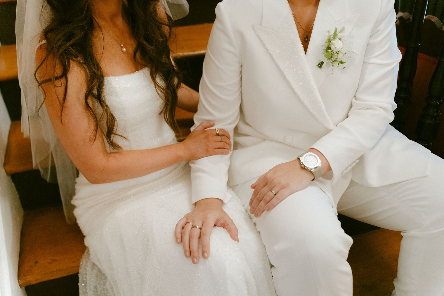 Close-up of two brides in white outfits showing wedding rings while seated together.