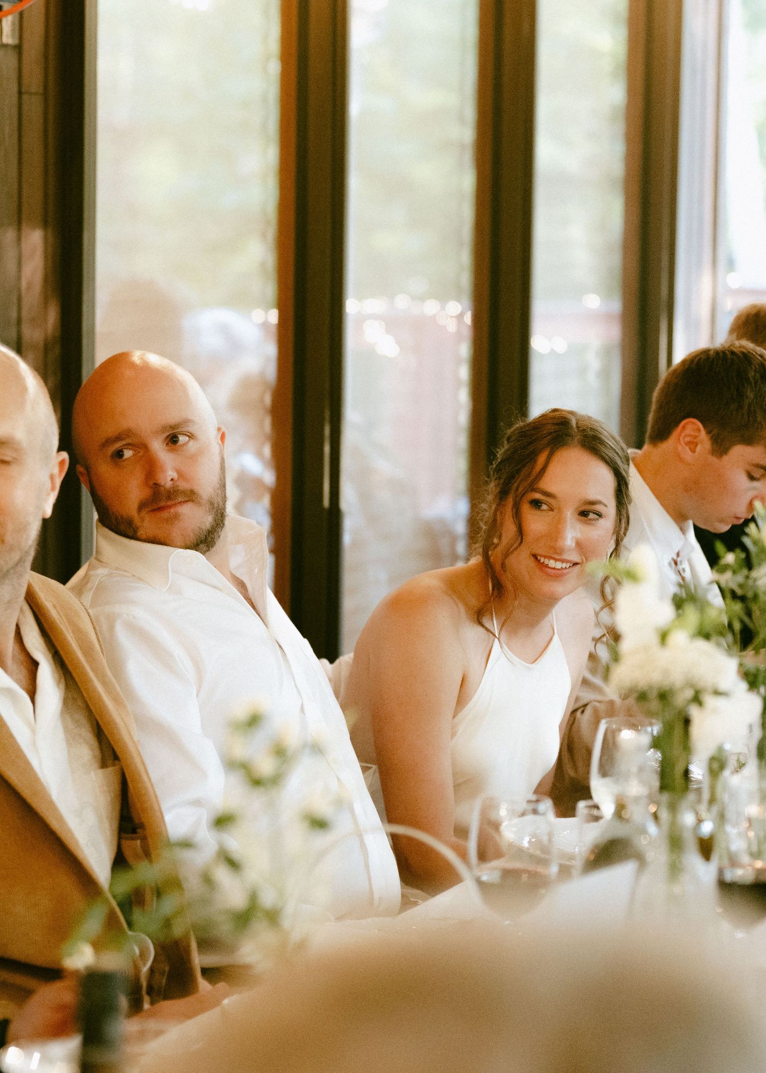 Bride in white dress and groom in white shirt seated at wedding reception table with floral centerpieces.