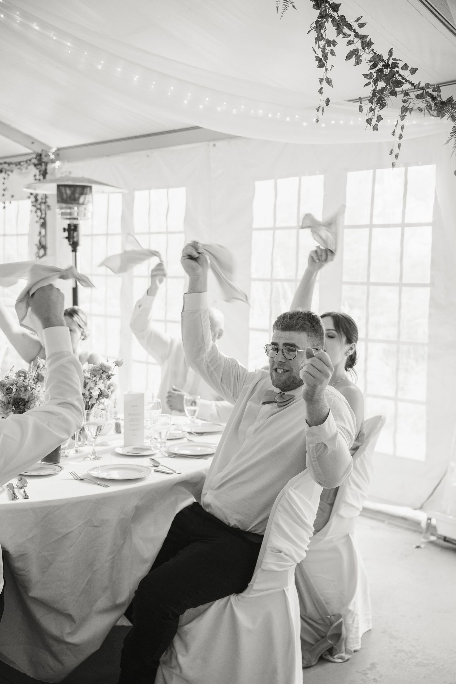 Black and white photo of wedding guests waving napkins in celebration at indoor reception.