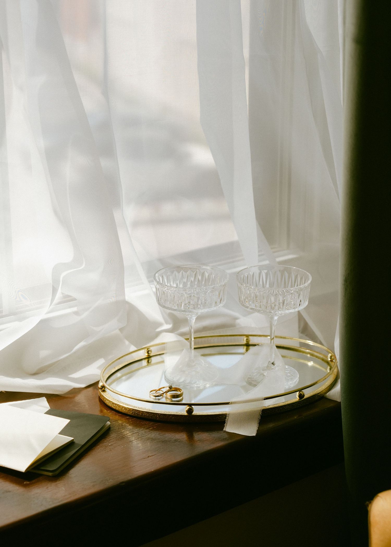 Wedding rings and crystal champagne coupes displayed on a gold tray by a sheer window.