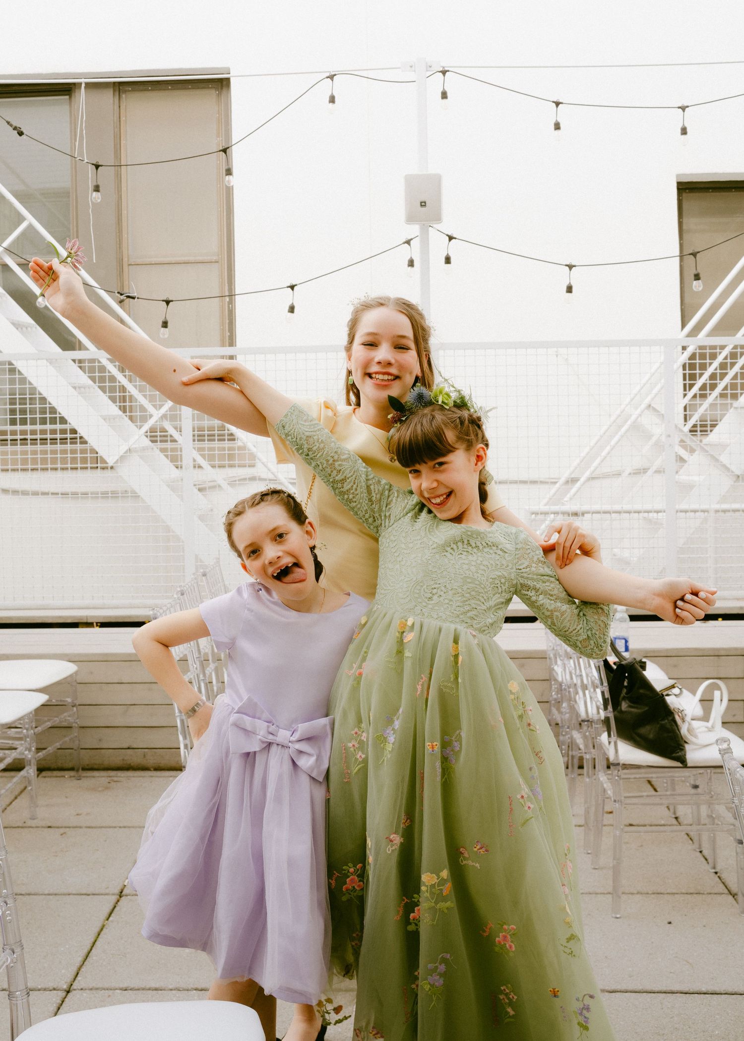 Three young girls in colorful dresses posing playfully at outdoor wedding venue with string lights.