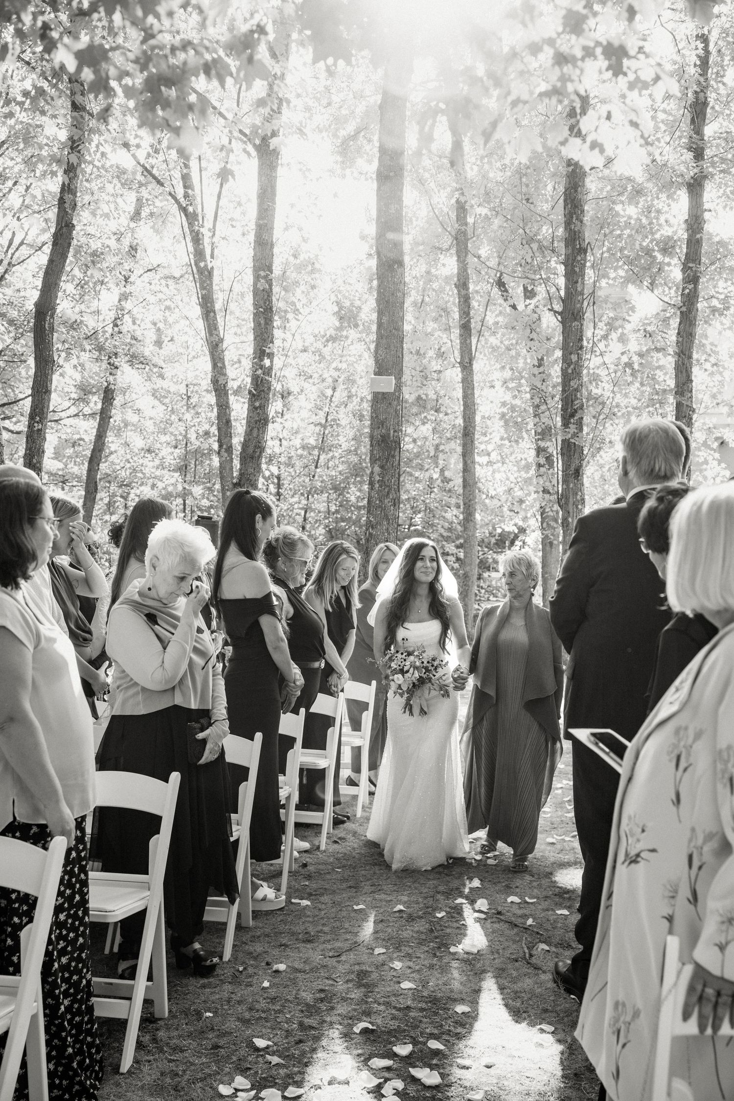 Black and white photo of a bride walking down the aisle at an outdoor forest wedding ceremony.