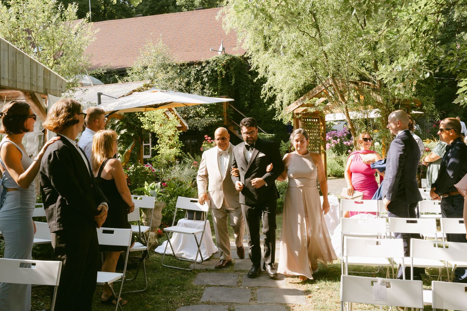 Bride walks down outdoor aisle with escort past seated wedding guests in garden ceremony.