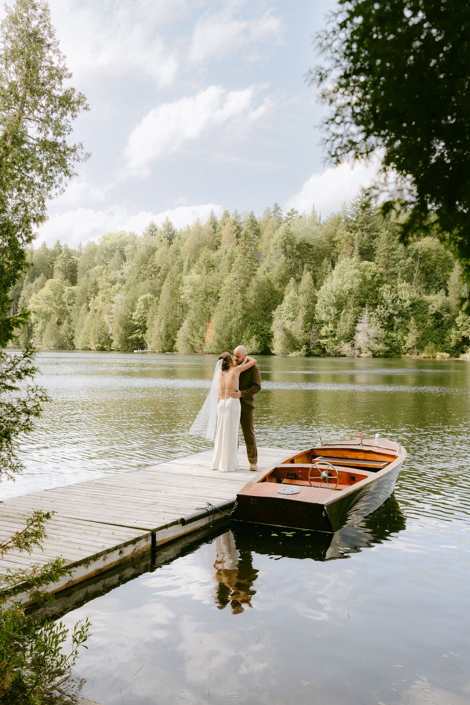 Bride and groom kissing on a lakeside dock beside a classic wooden boat with forest reflection in calm water.