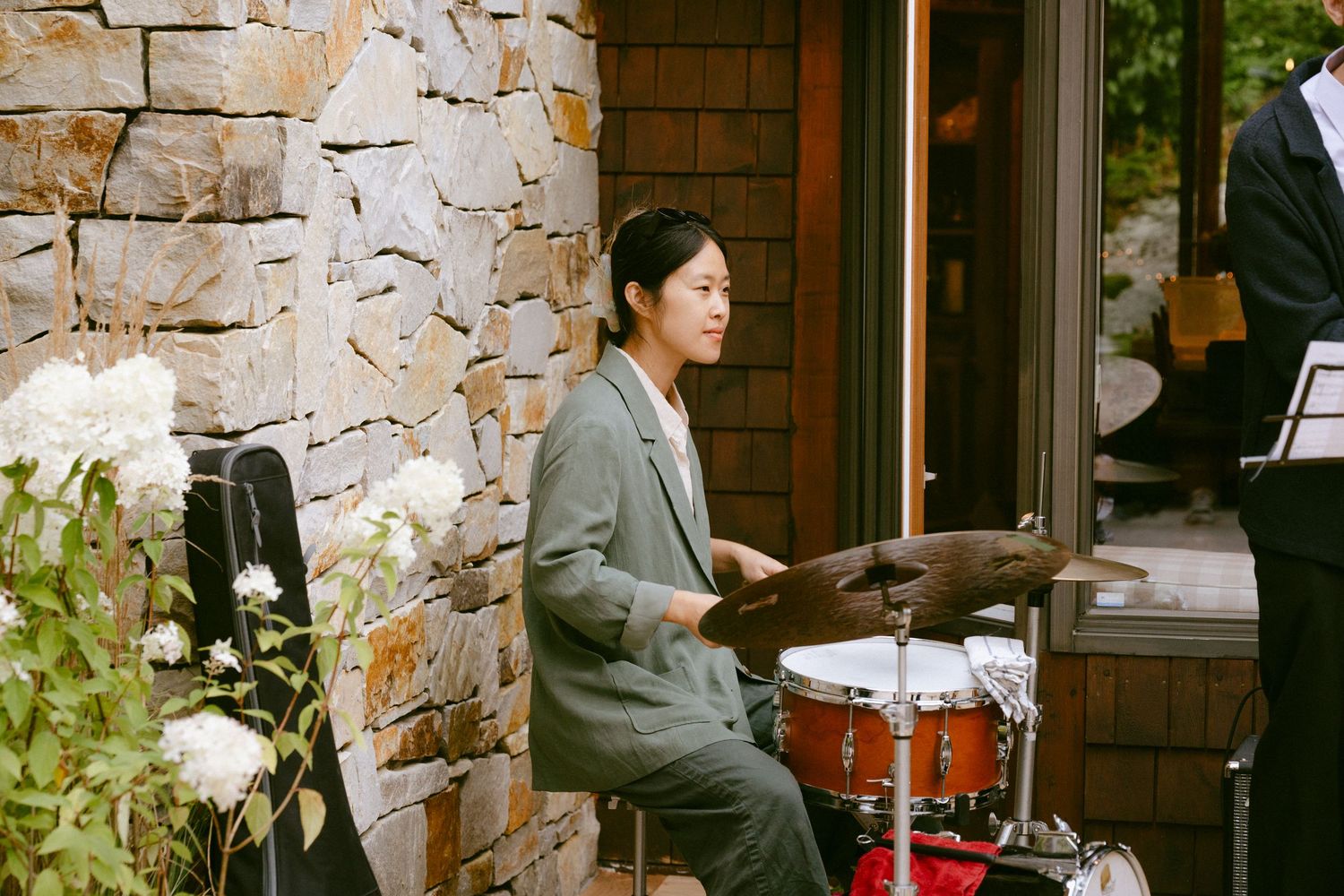 Female drummer in grey suit performing outdoors at wedding reception near stone wall with flowers.