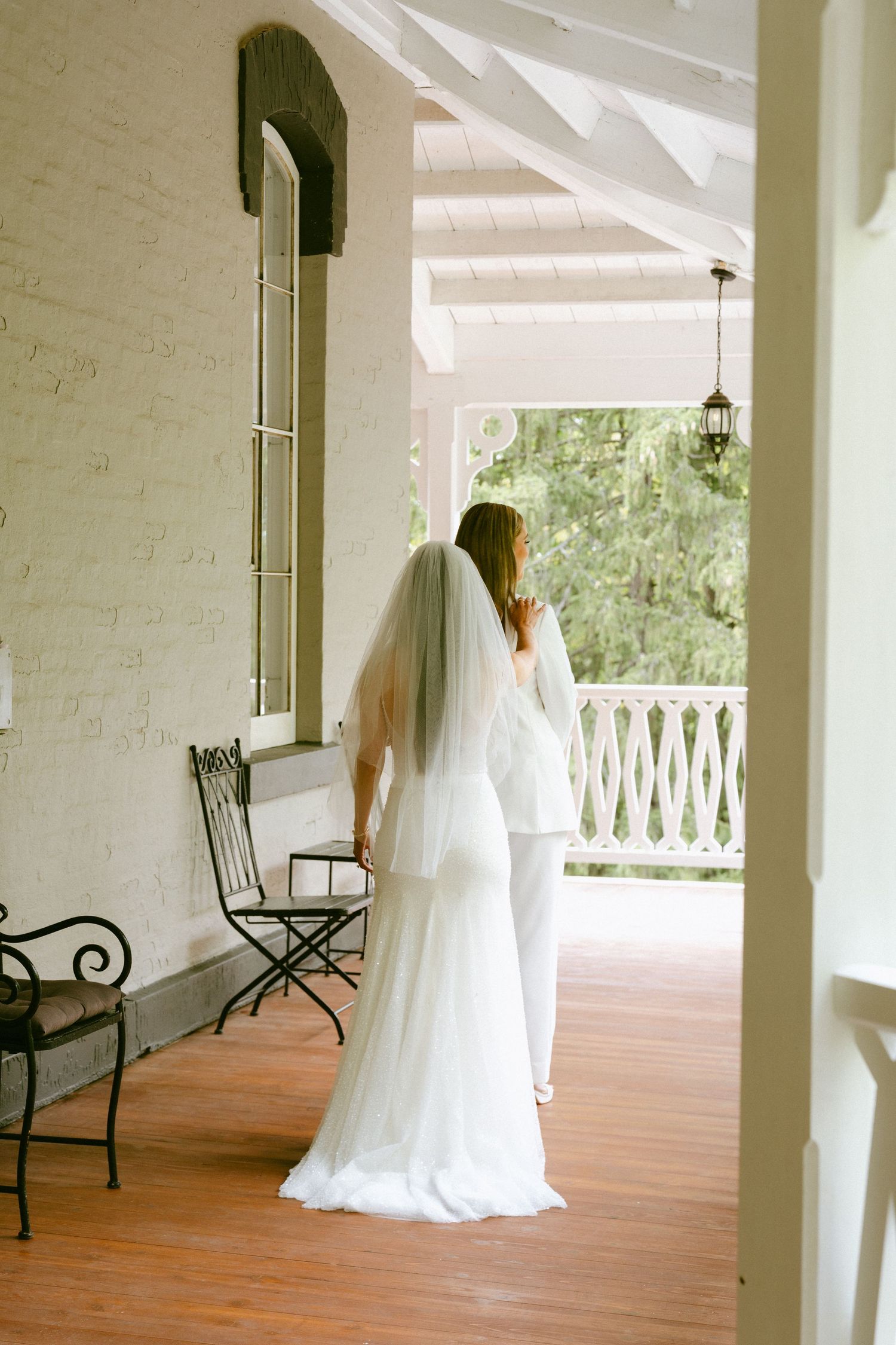 Bride in white gown and veil stands on elegant porch of white brick venue.