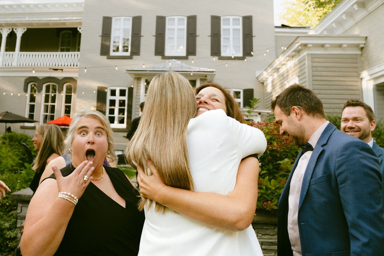 Bride in white dress hugging a guest while others react with joy at outdoor wedding reception.