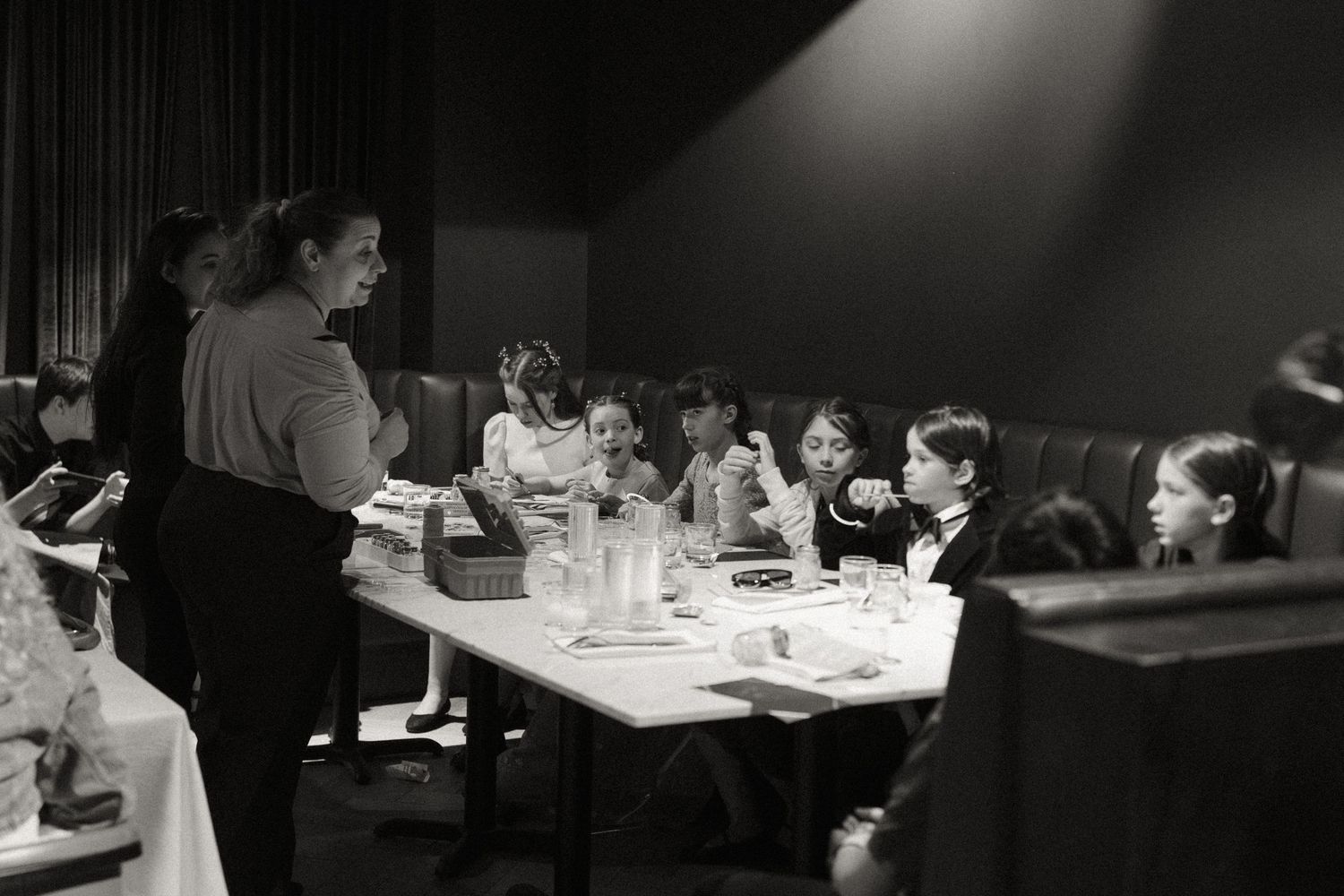 Teacher with children seated at a long table during an indoor dining event in black and white.