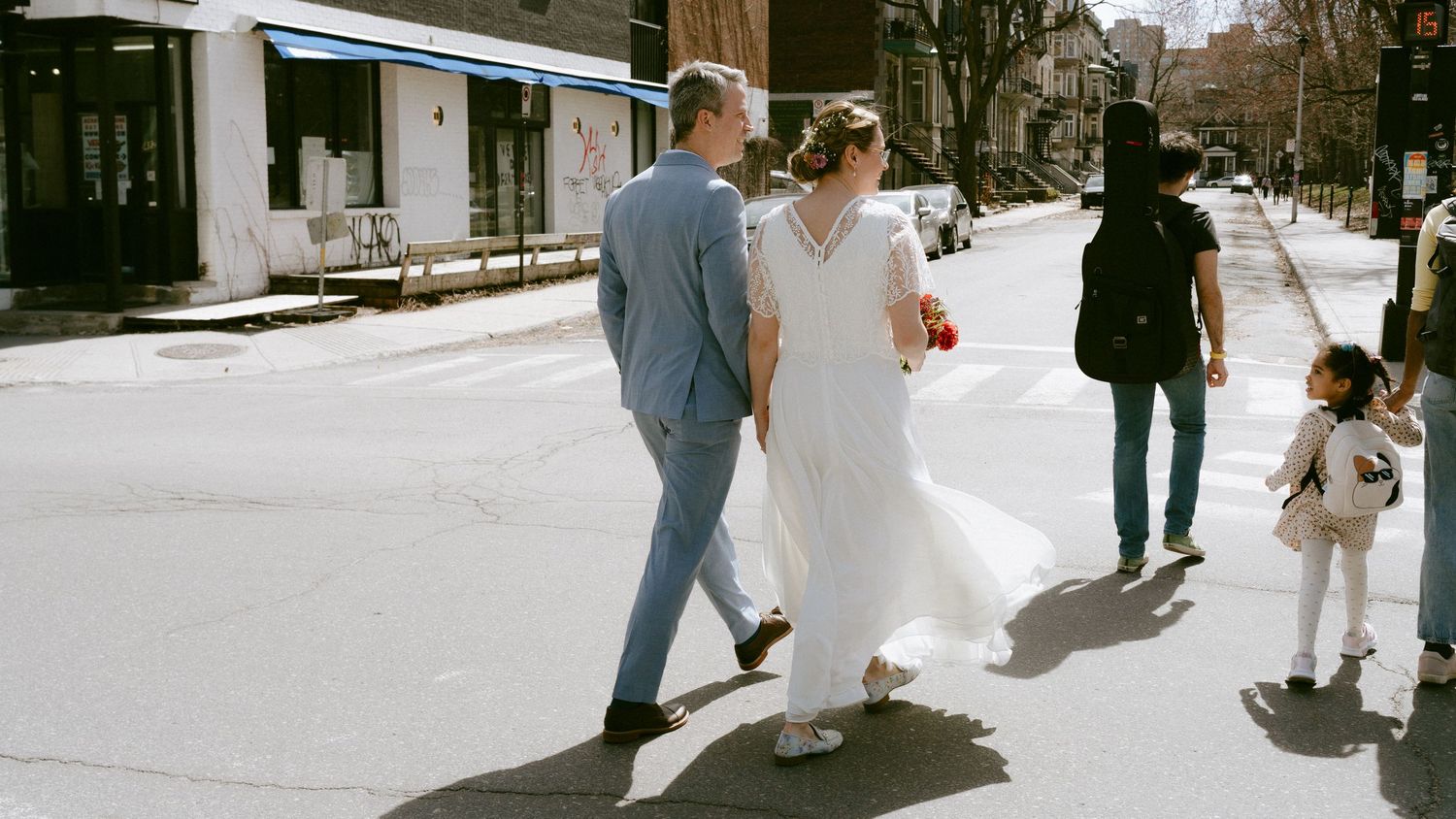 Bride and groom walk hand in hand down a sunny city street on their wedding day.