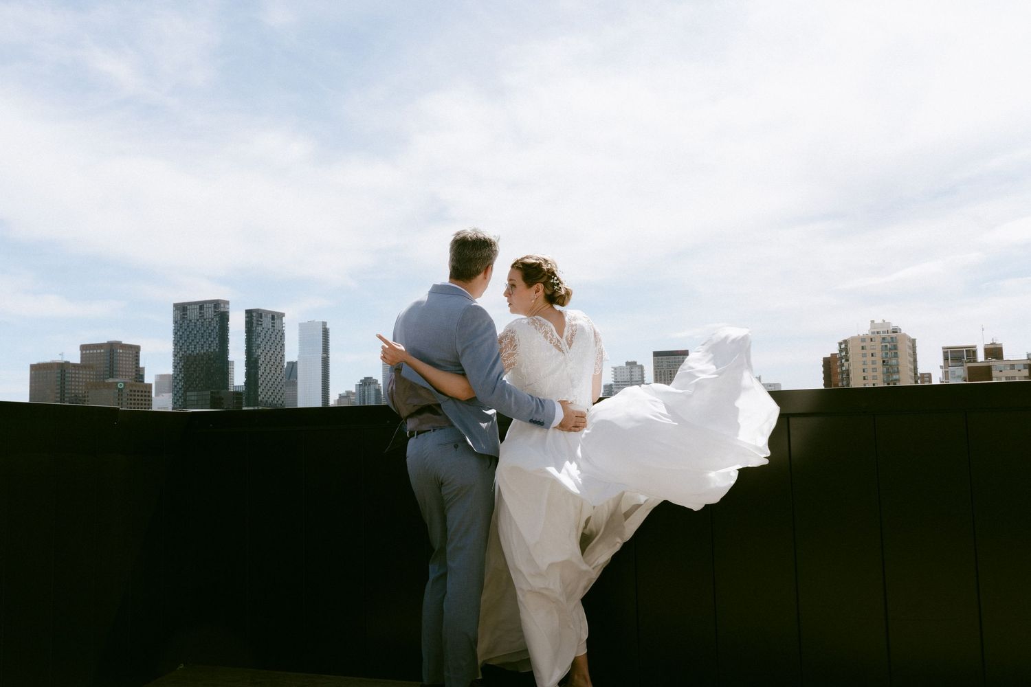 Bride's dress billows in the wind as couple embraces on a rooftop with city skyline.