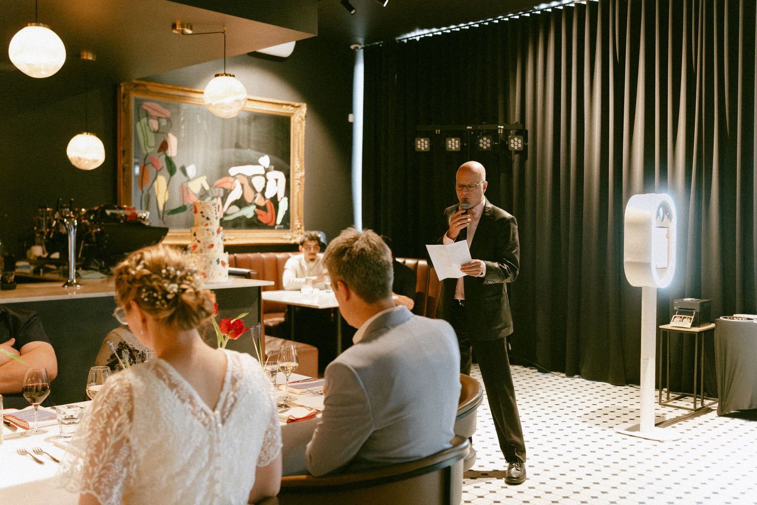 Wedding MC speaking at a microphone while bride and groom sit at their table in a stylish dark restaurant.