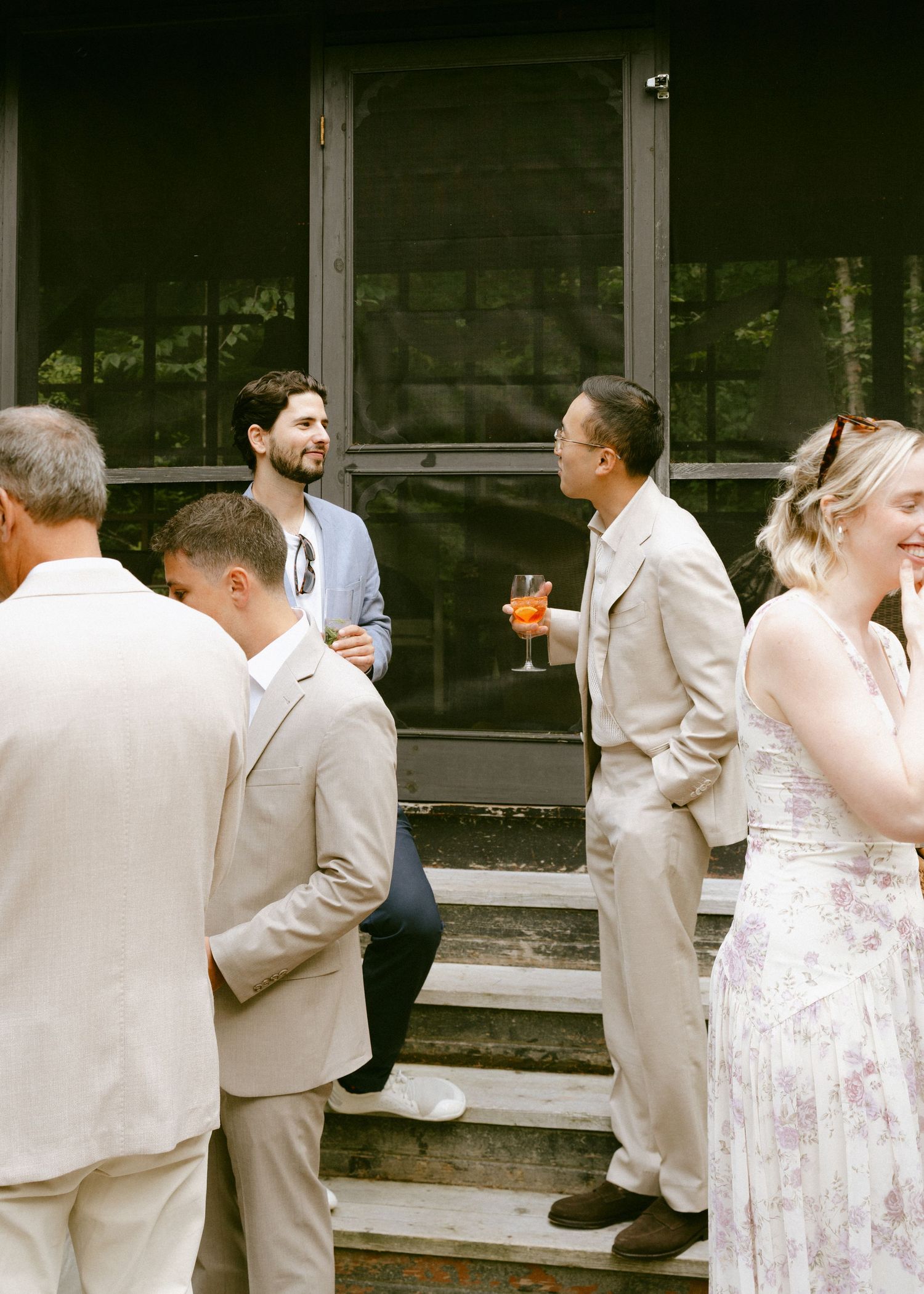 Wedding guests in elegant attire socializing on steps, man holding cocktail at outdoor reception.