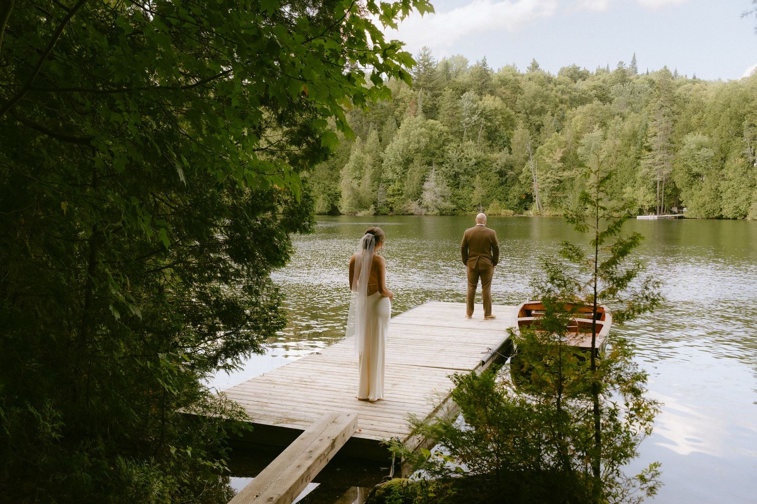 Bride and groom standing apart on a wooden lakeside dock surrounded by lush green forest trees.