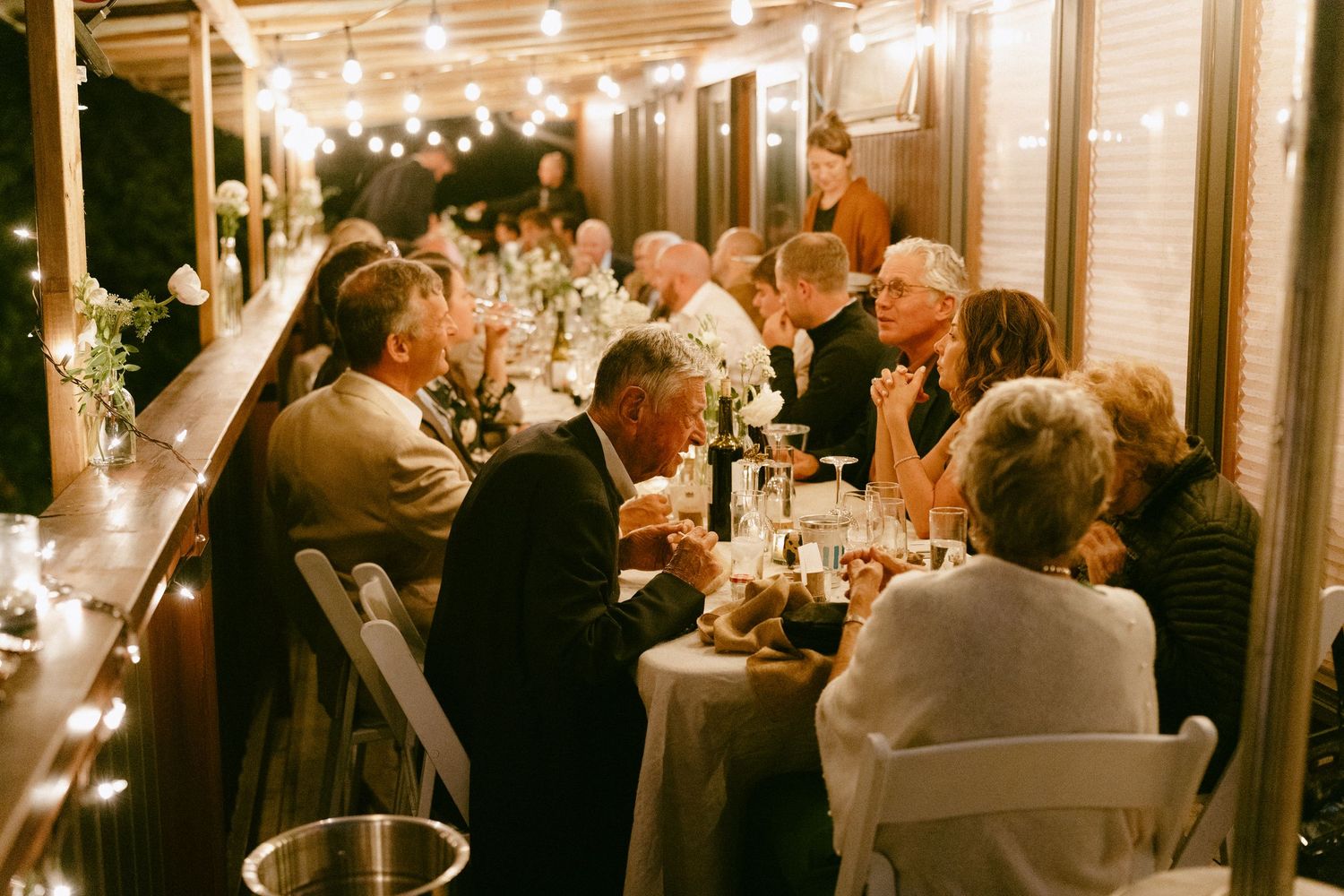 Guests seated at long wedding reception table adorned with string lights and floral centerpieces.