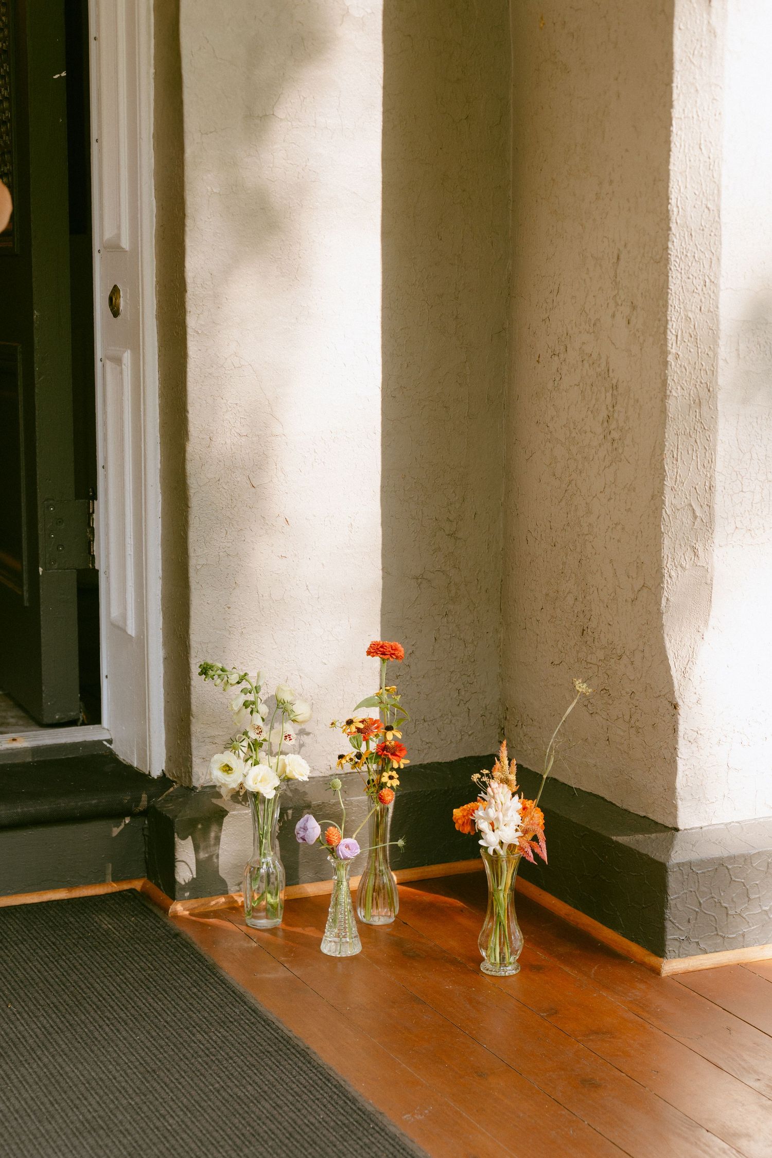 Four glass bud vases with colorful wildflowers arranged on a wooden porch floor by a doorway.