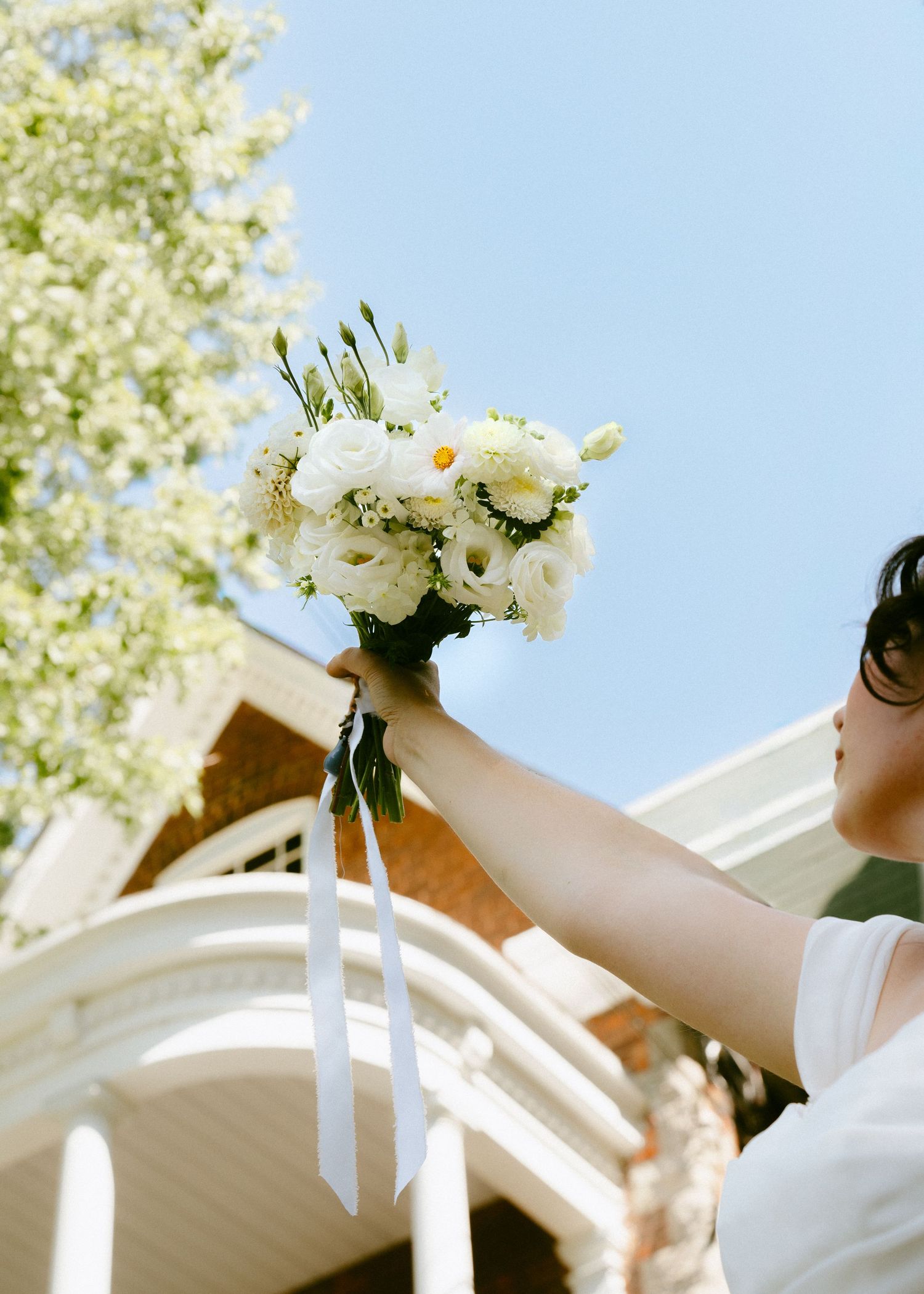 Bride holds white floral bouquet with ribbon streamers up against a bright blue sky.