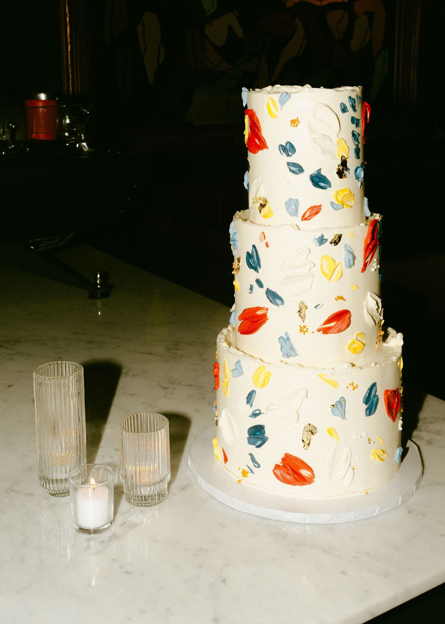 Three-tier white wedding cake decorated with colorful painted brushstroke details in red, blue, and yellow hues.