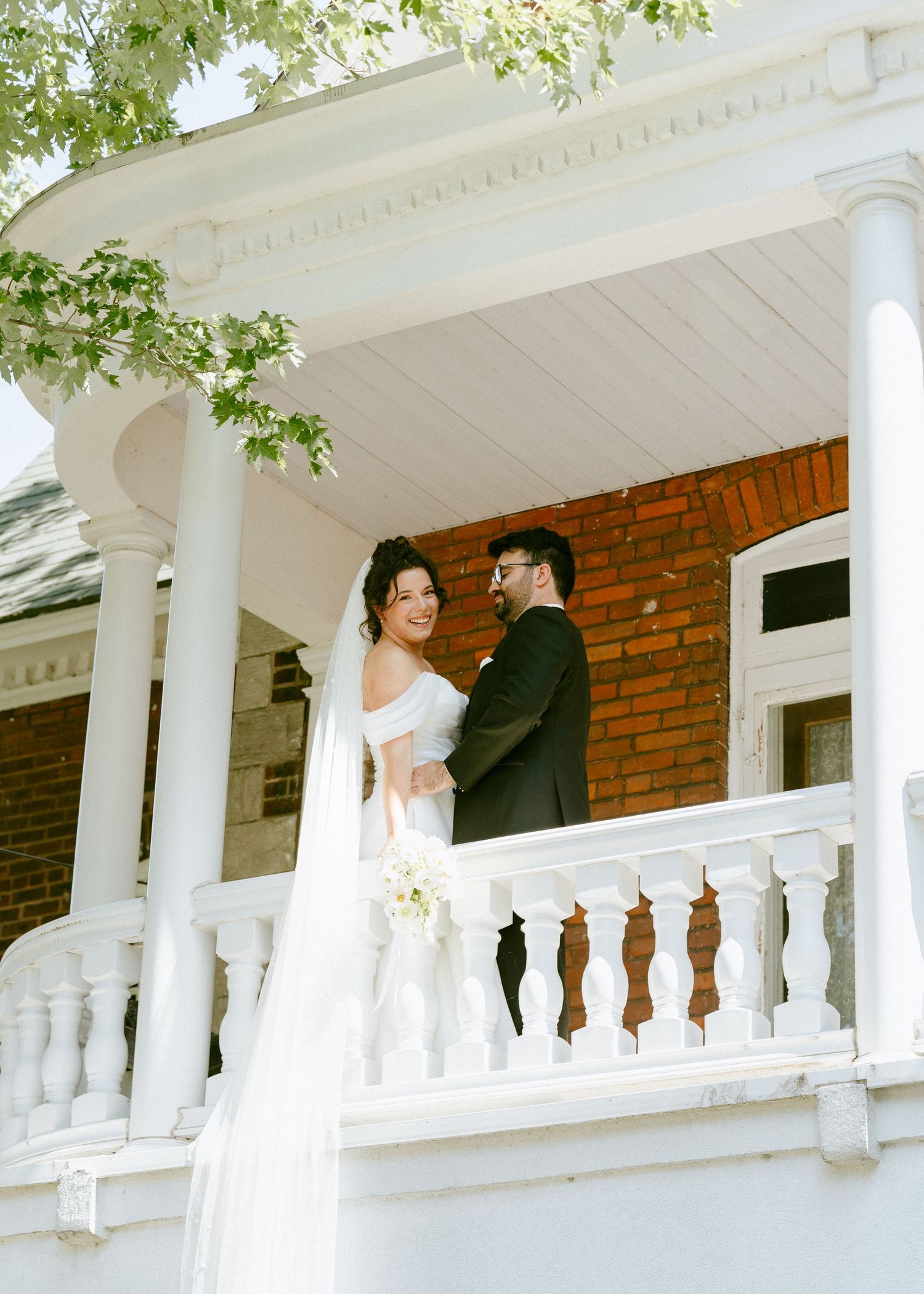 Bride and groom stand together on white Victorian porch balcony of brick wedding venue.