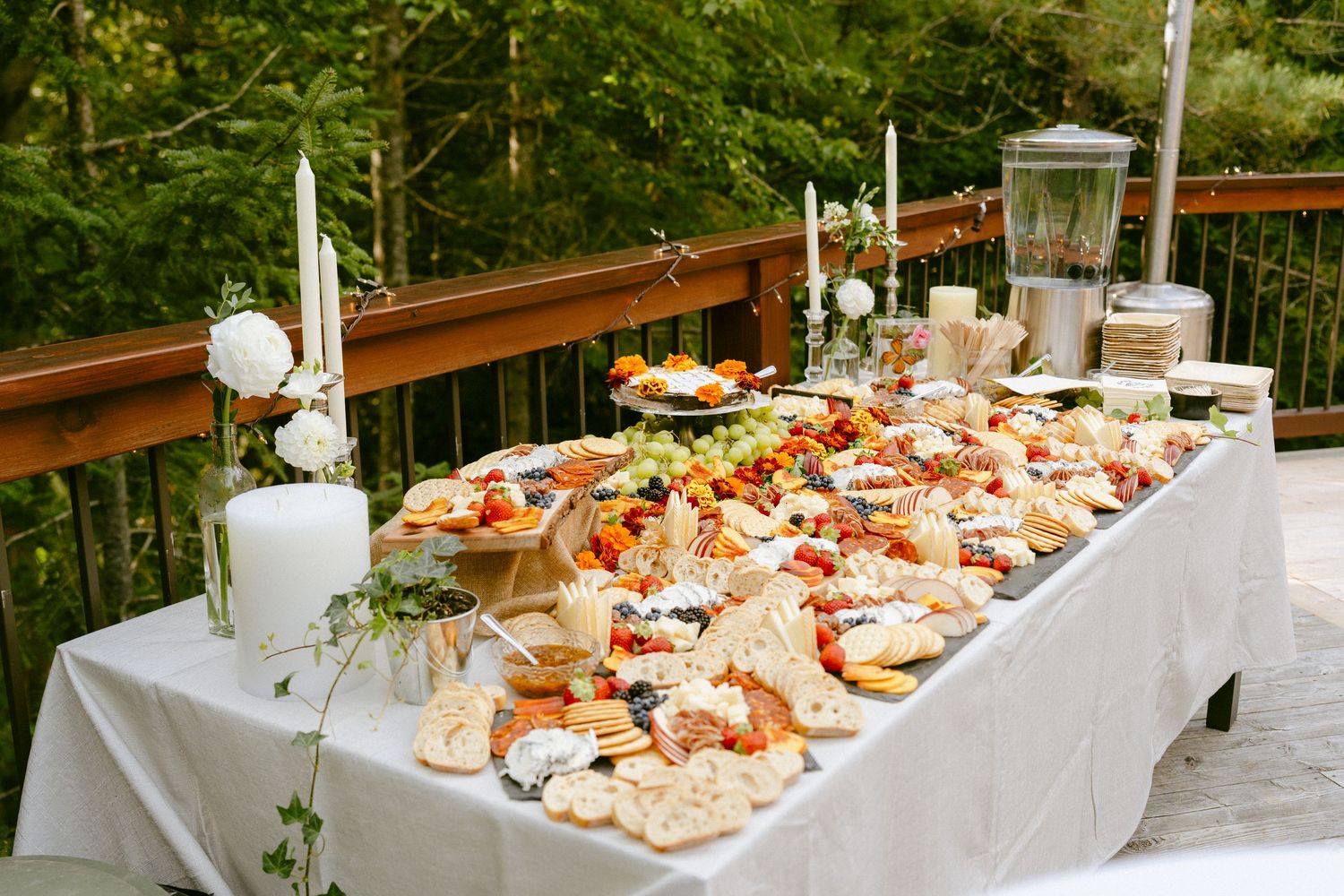 Full outdoor wedding grazing table spread with charcuterie, fruits, cheeses, and candles on a forest deck.