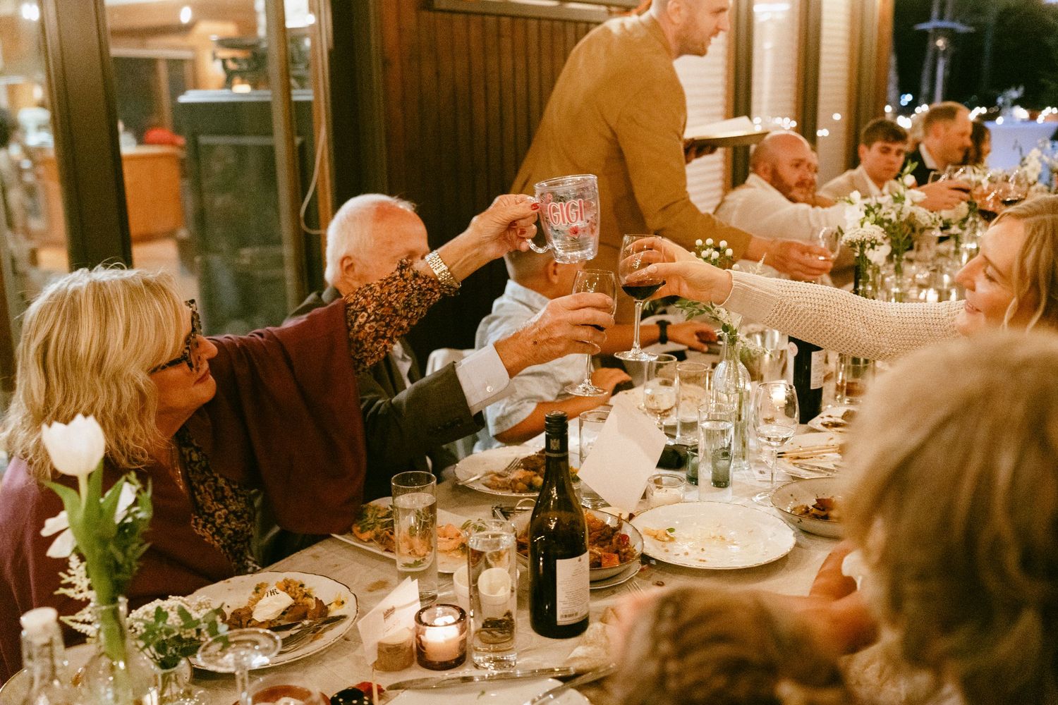 Wedding guests raising glasses in a toast at a candlelit reception dinner table.
