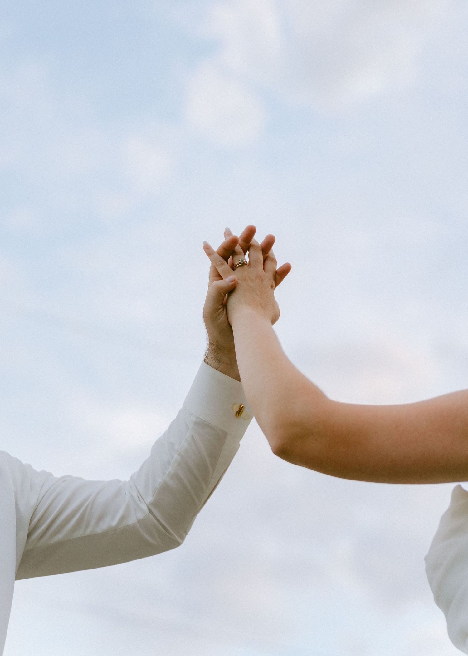 Bride and groom holding hands raised against a cloudy sky.