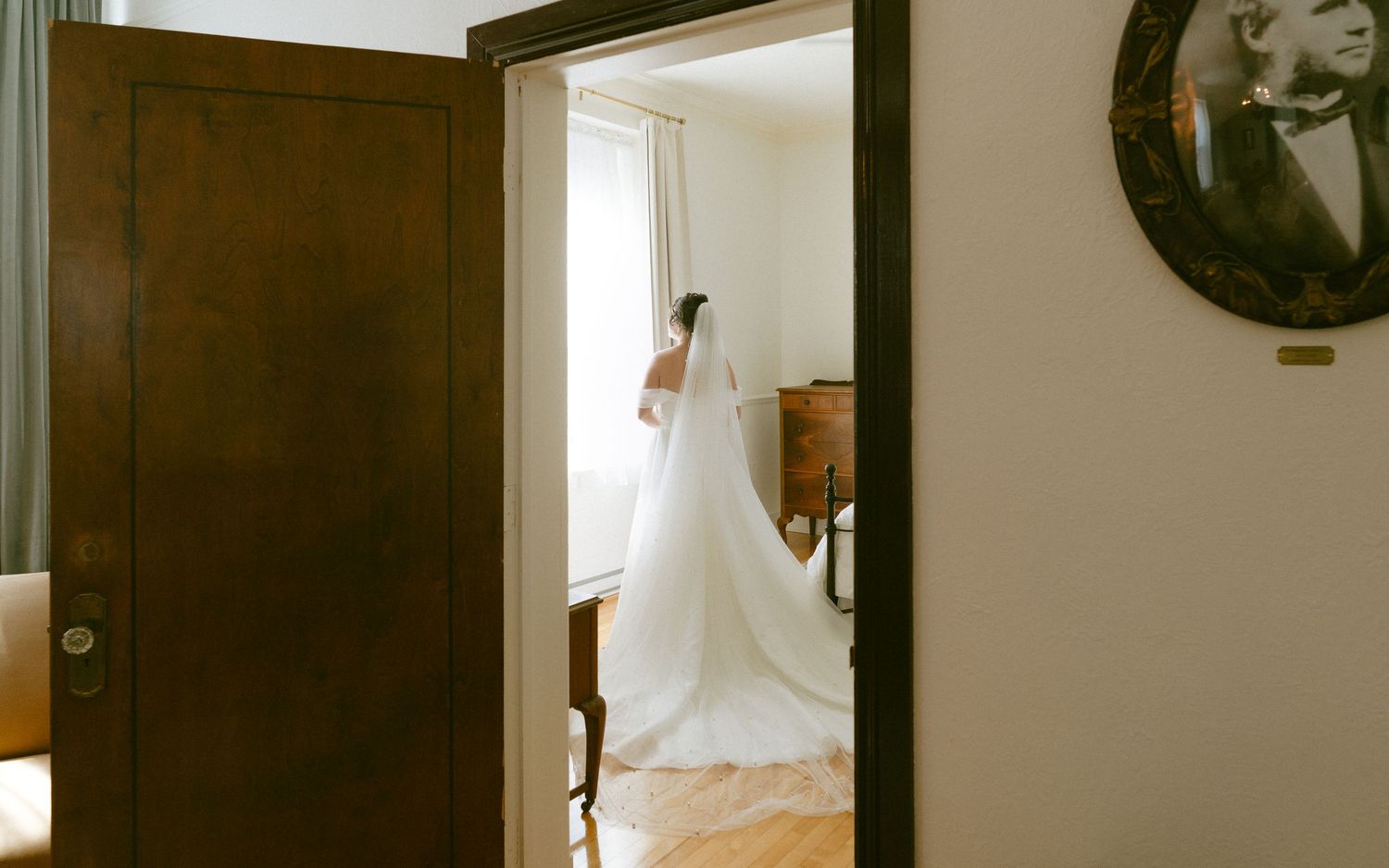 Elegant bride in long white wedding gown and veil seen through dark wooden doorway in vintage room.