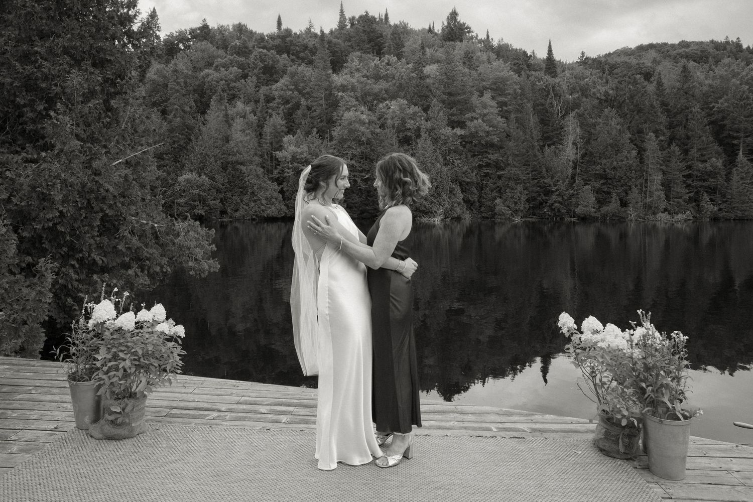 Two brides sharing an intimate moment on a lakeside dock with lush forest backdrop in black and white.