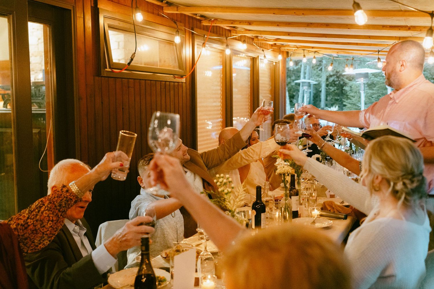 Wedding guests raising glasses in a toast under warm string lights at an indoor dinner.