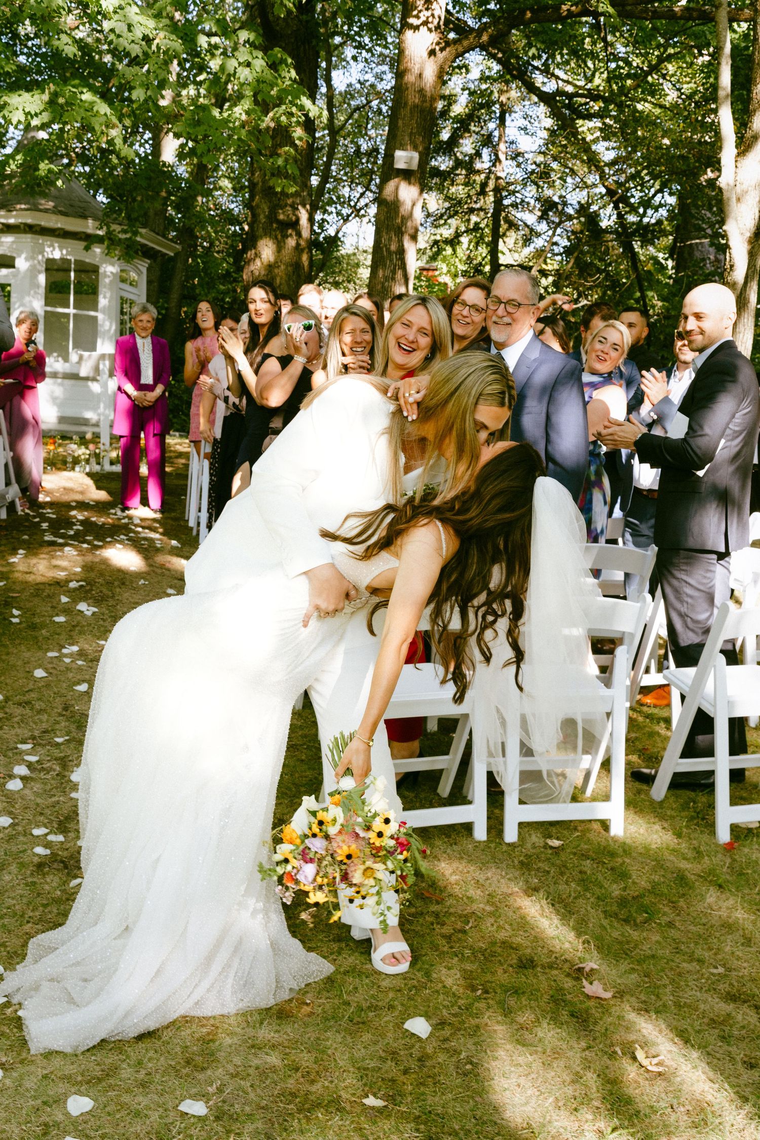 A bride in a white gown dips joyfully while holding a colorful bouquet during her wedding ceremony.