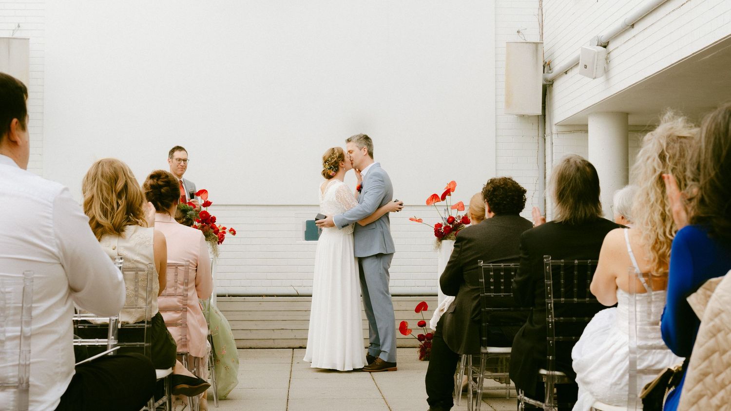 Couple sharing their first kiss during an intimate small wedding ceremony with guests holding red floral bouquets.