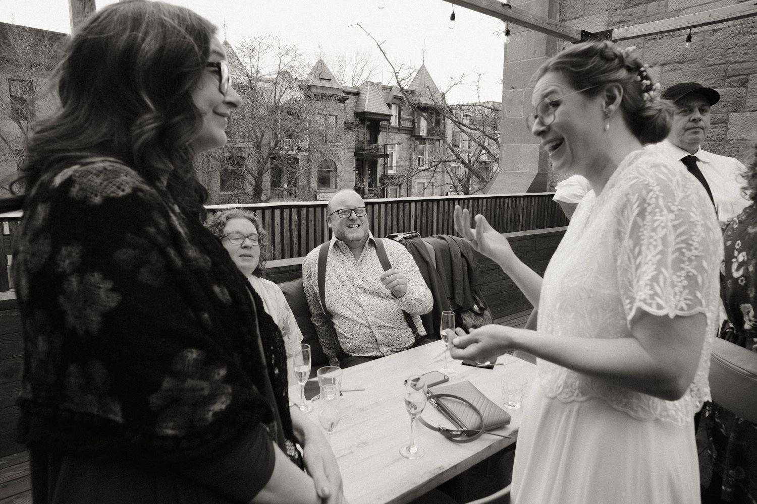 Bride in white lace dress laughs with guests at outdoor rooftop wedding reception.