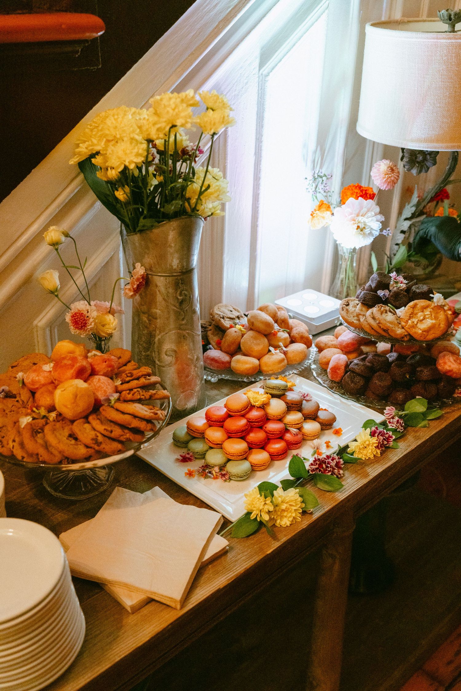 Elegant wedding dessert table featuring colorful macarons, cookies, and fresh floral arrangements by a window.