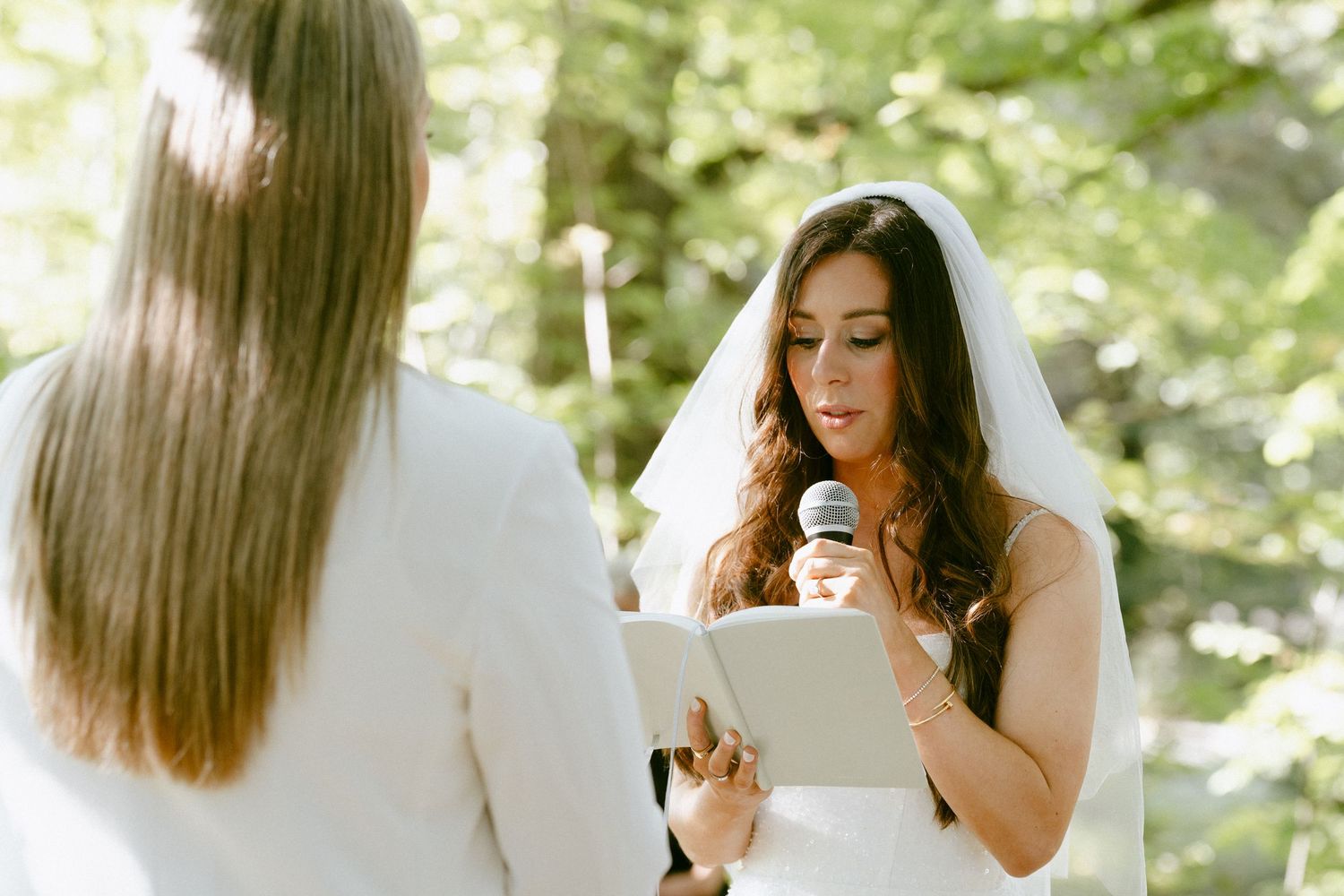 Bride in veil reading wedding vows from a book while holding a microphone outdoors.
