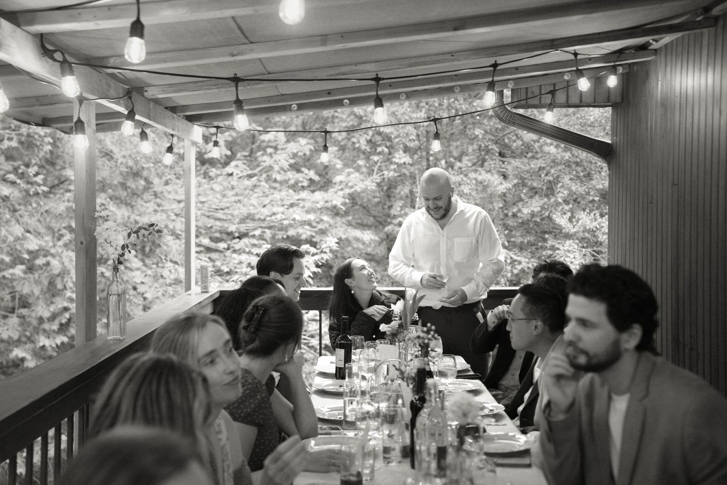 Guests gather at a long outdoor dinner table under string lights as a server attends to them.