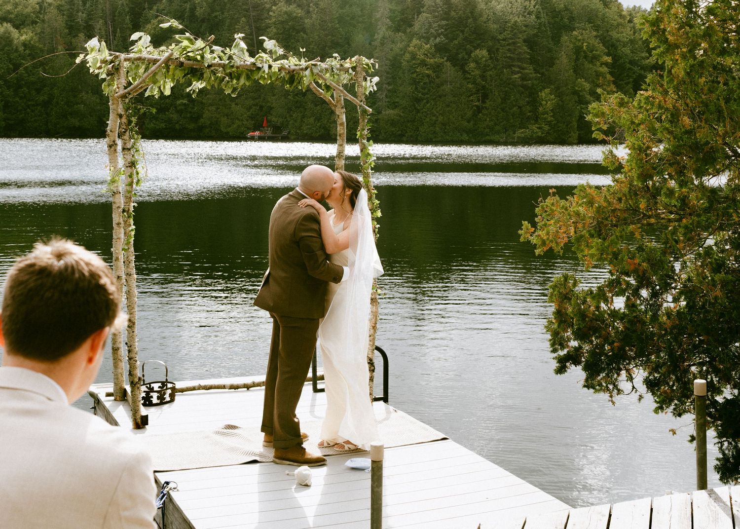 Bride and groom share their first kiss under a floral arch on a peaceful lakeside dock.