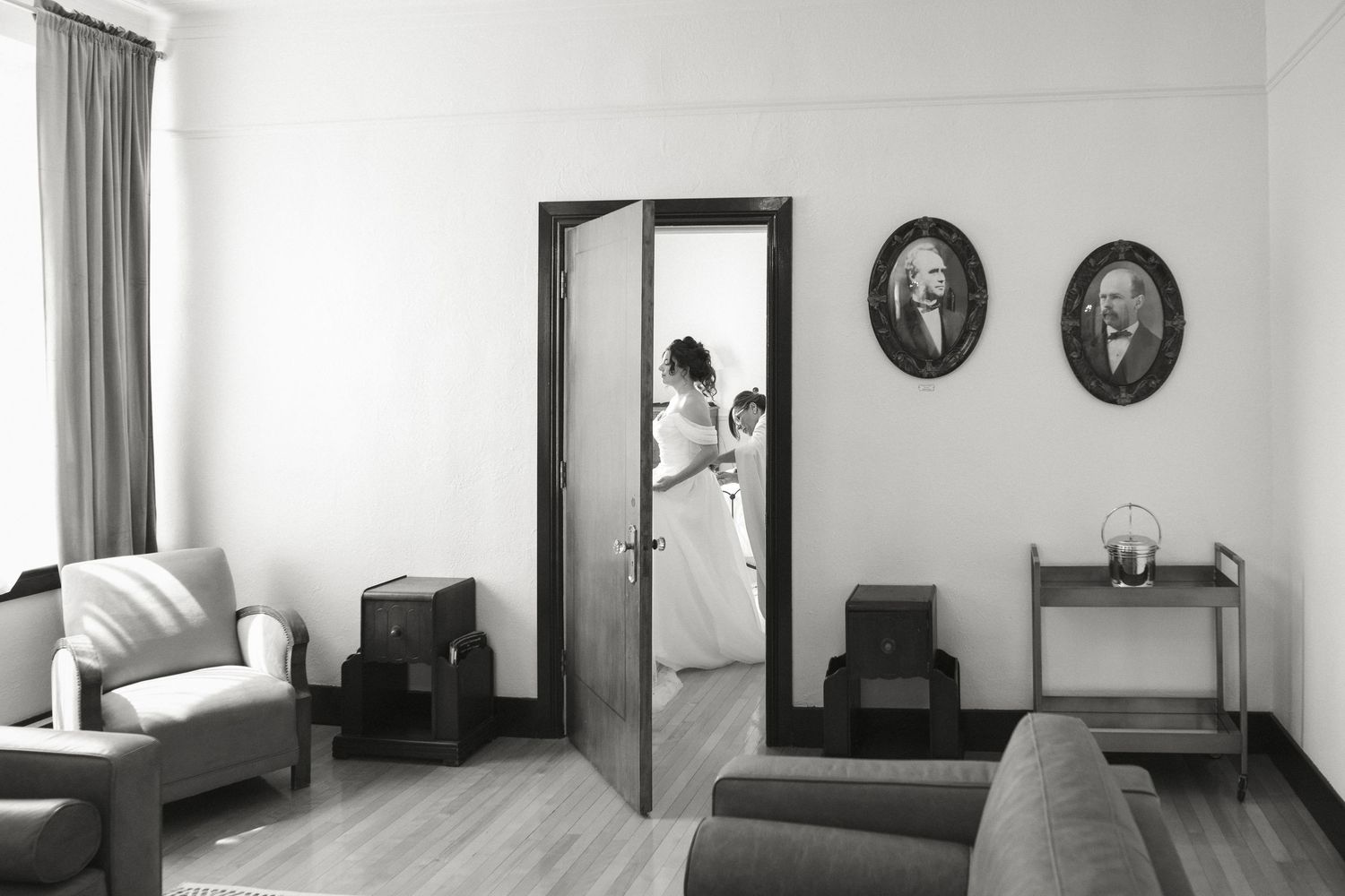 Bride in wedding gown visible through open double doors, reflected in mirror, black and white portrait.