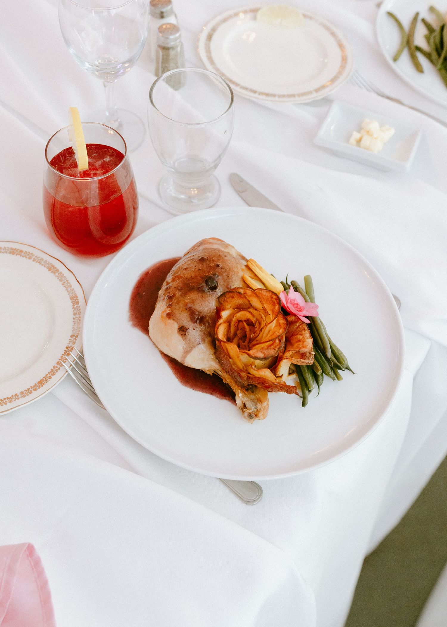 Wedding reception dinner plate with roasted chicken, green beans, and gravy beside red cocktail.