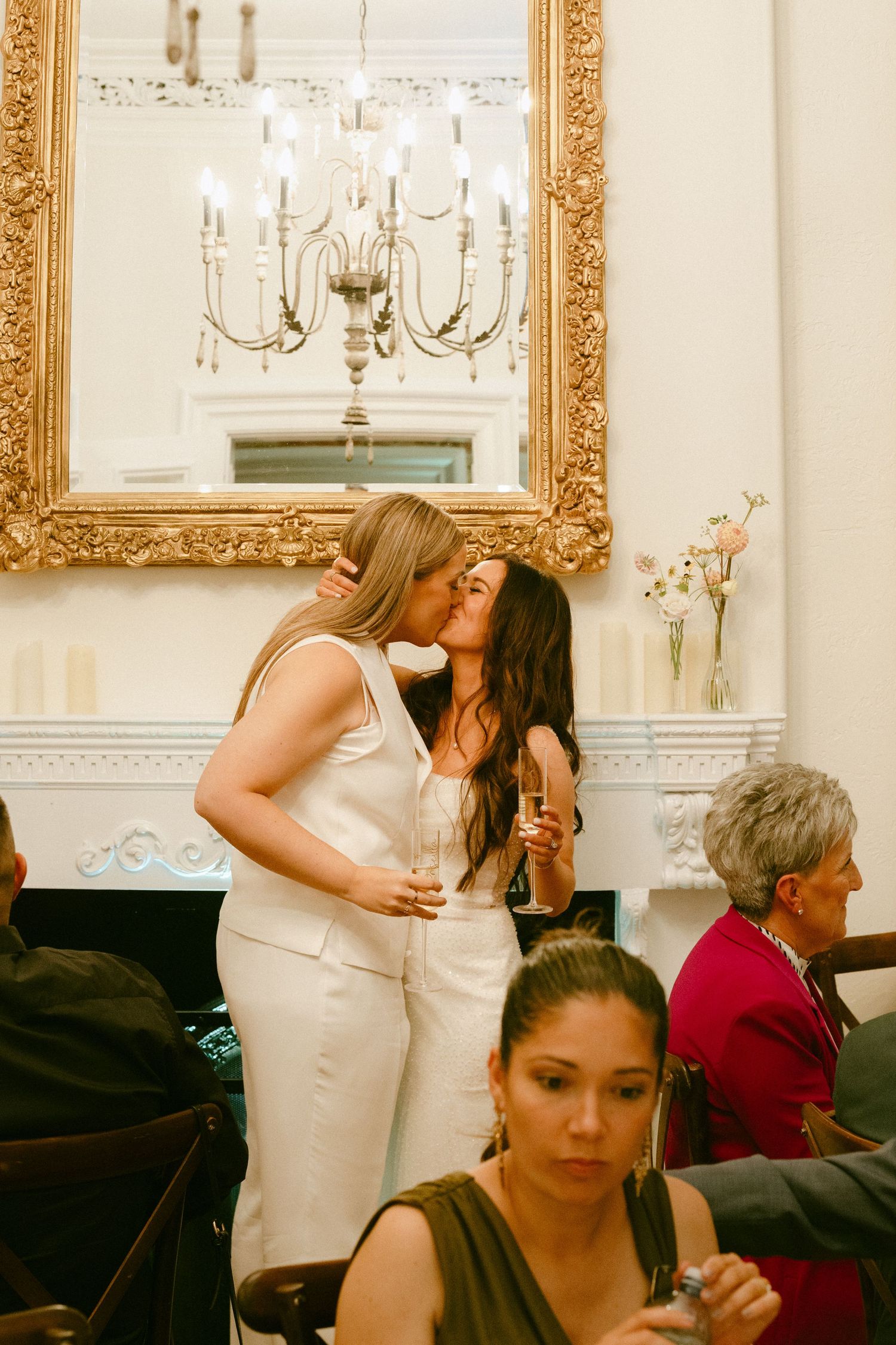 Two brides sharing a kiss at their wedding reception beneath a gold-framed mirror and chandelier.